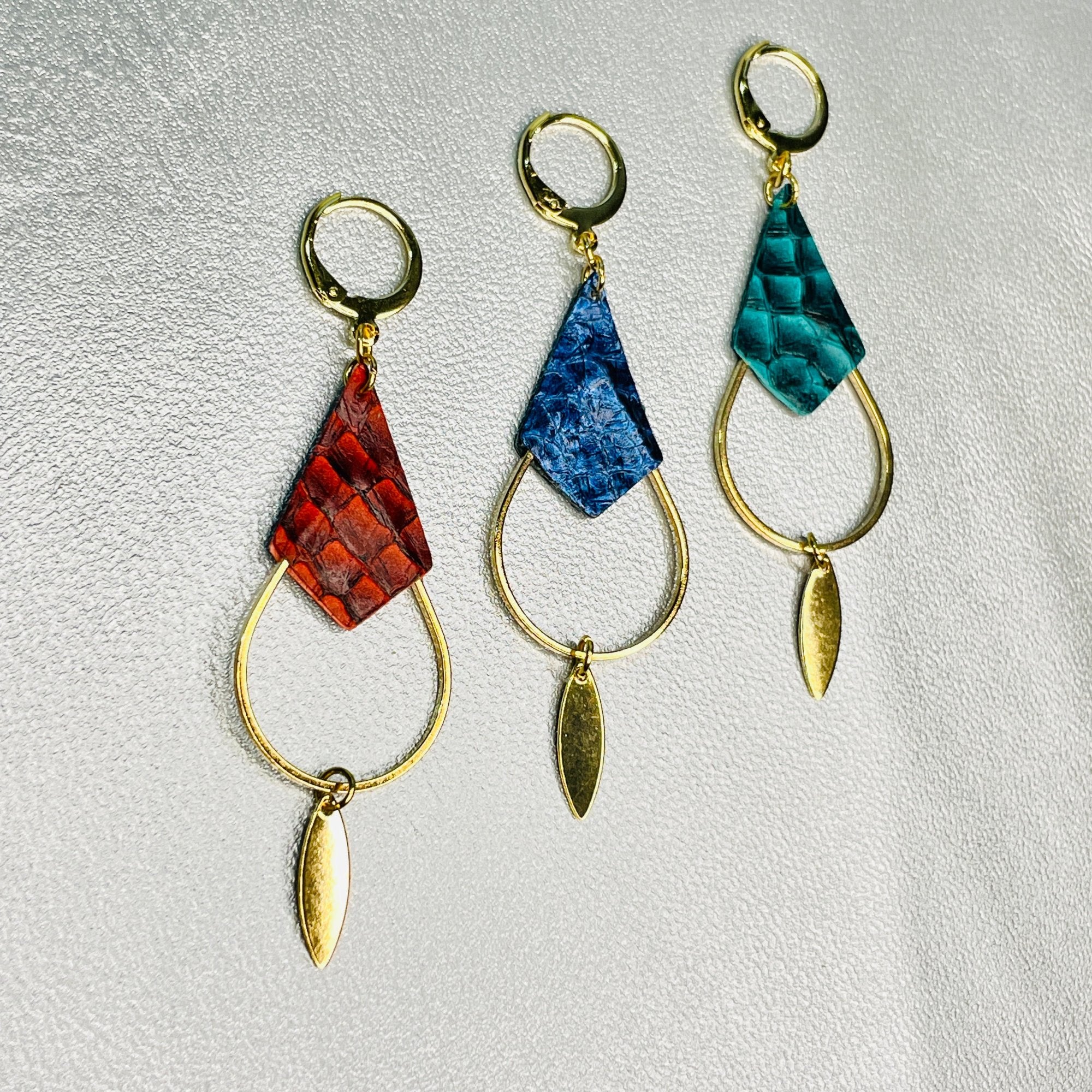 Boucles d'oreilles Gali de Nune, dorées à l'or fin avec triangles de cuirs marins, cuirs de poisson saumon rouge carmin écaille, bleu saphyr métallisé et vert émeraude écaille, de forme grande goutte, sur fond cuir gris argenté