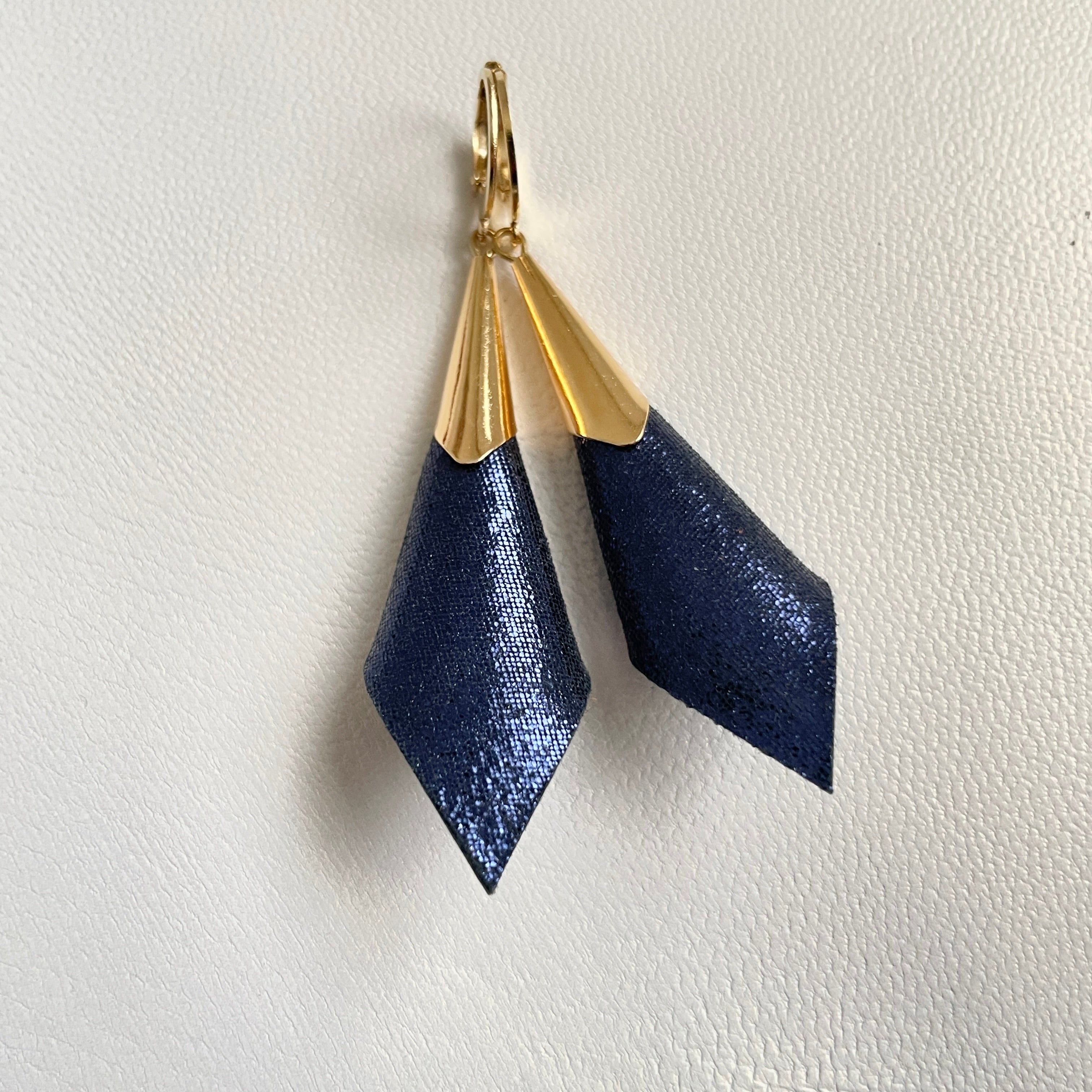 Boucles d'oreilles dorées Nune modèle Alice en forme de cônes de cuir bleu marin pailleté, sur fond cuir blanc cassé nacré