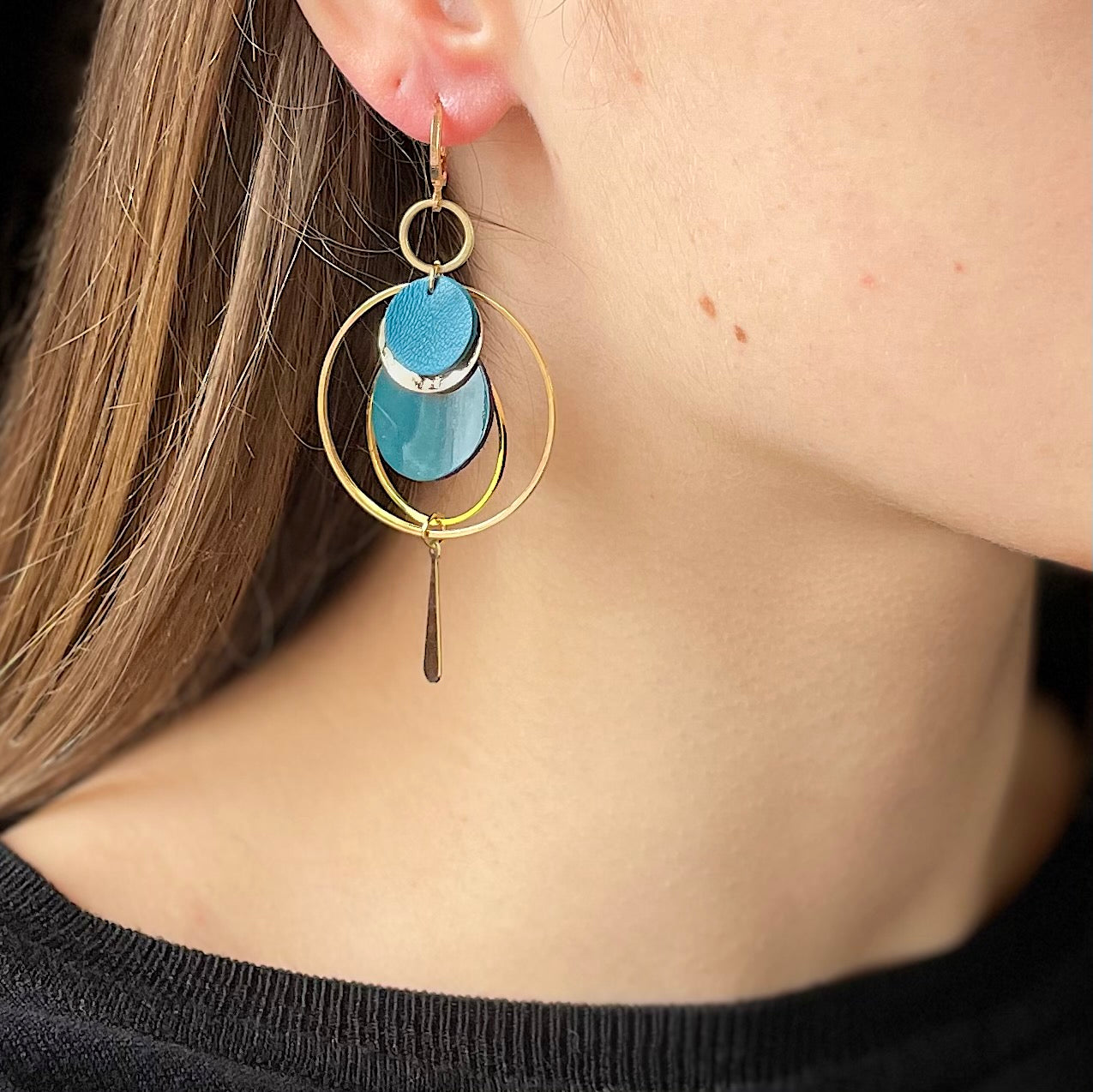 Boucle d'oreille dorée Nune modèle Galassia en cuirs bleu canard satiné et bleu canard vernis portée par un modèle aux cheveux blonds et au teint pâle