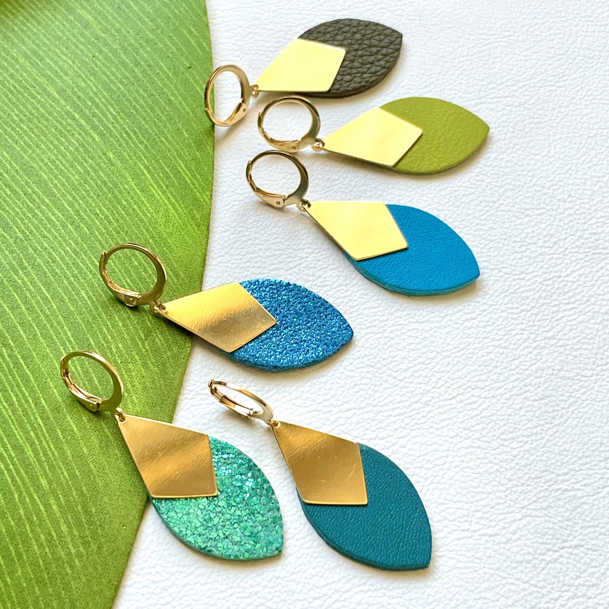 Gamme boucles d'oreilles dorées Nune en forme d'olives modèle Olivia en cuirs recyclés dans les tons bleus et verts sur fond blanc et vert