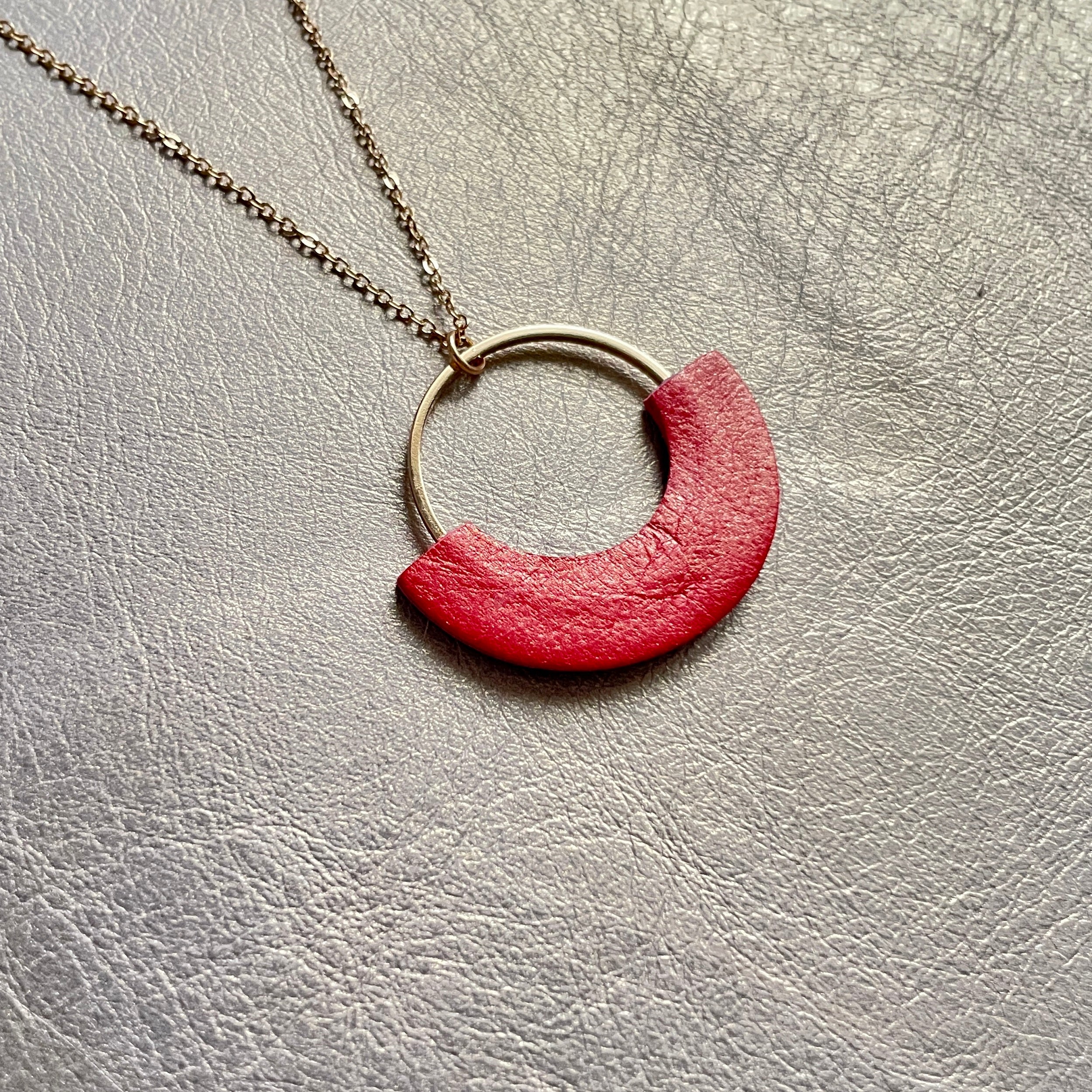 Collier fin Nune modèle Mezzaluna doré avec pendentif rond en forme de demi-lune en cuir recyclé coquelicot nacré, vue de biais, sur fond beige doré