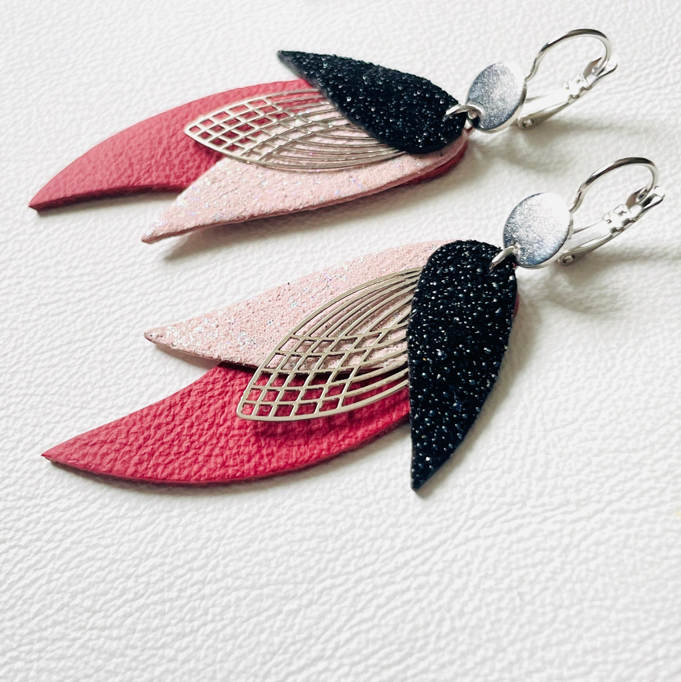 Boucles d'oreilles argentées Nune en forme de plumes, modèle Ara en cuirs noir grainé pailleté, rose poudré irisé et rouge foncé mat mat sur fond blanc