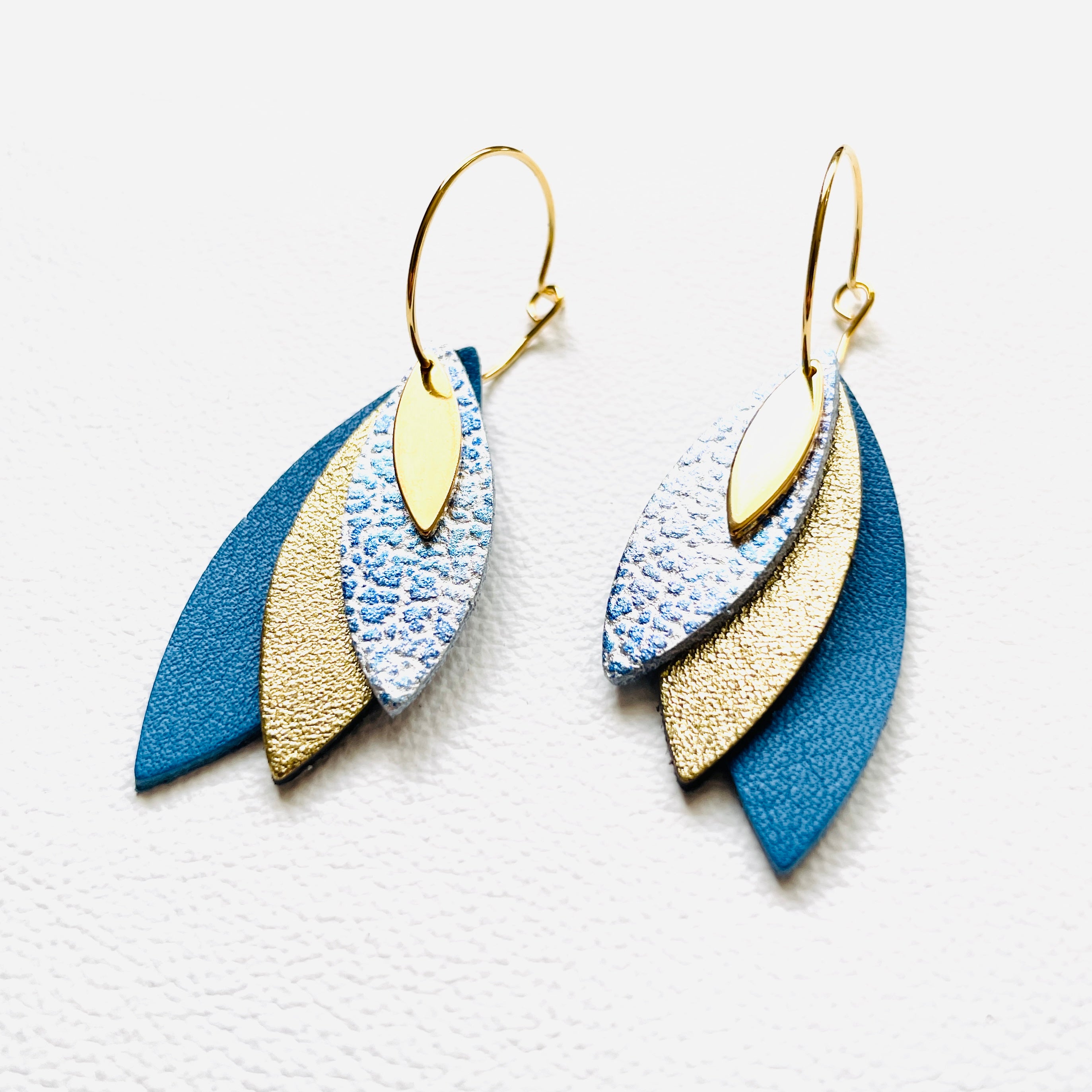 Boucles d'oreilles créoles dorées Nune modèle Piume en cuirs bleu ciel métal patiné, doré satiné et bleu denim mat sur fond blanc