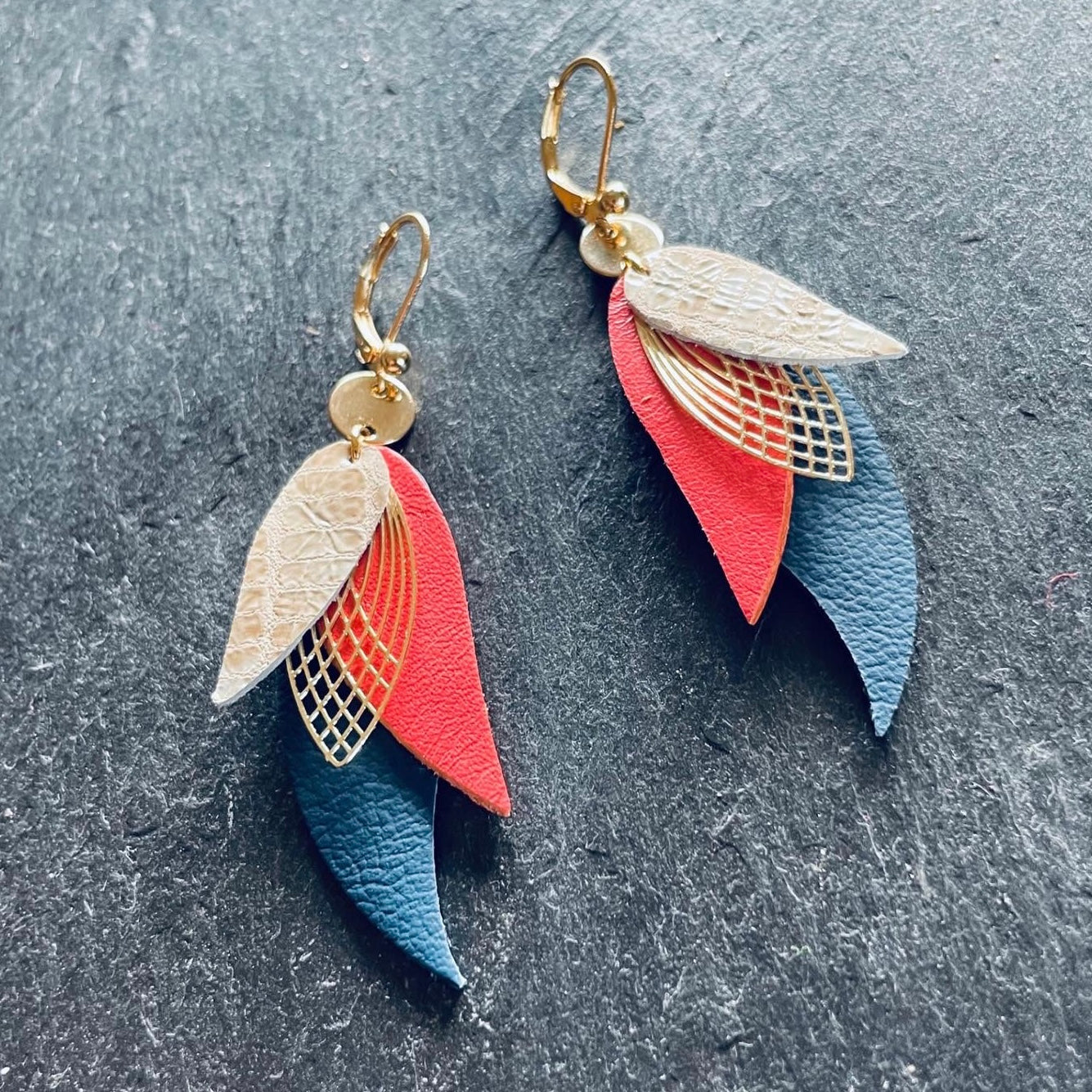 boucles d'oreilles dorées Nune modèle Ara en plumes de cuir gris façon serpent, rose saumon et bleu nuit mat sur fond ardoise