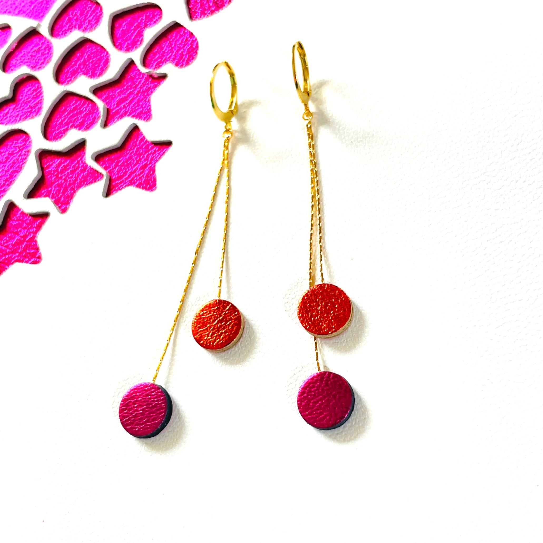 Longues et fines boucles d'oreilles Nune dorées terminées petits disques de cuir recyclé orange sanguine métallisé et fushia métallisé, sur fond blanc et fushia étoilé. Modèle Merry