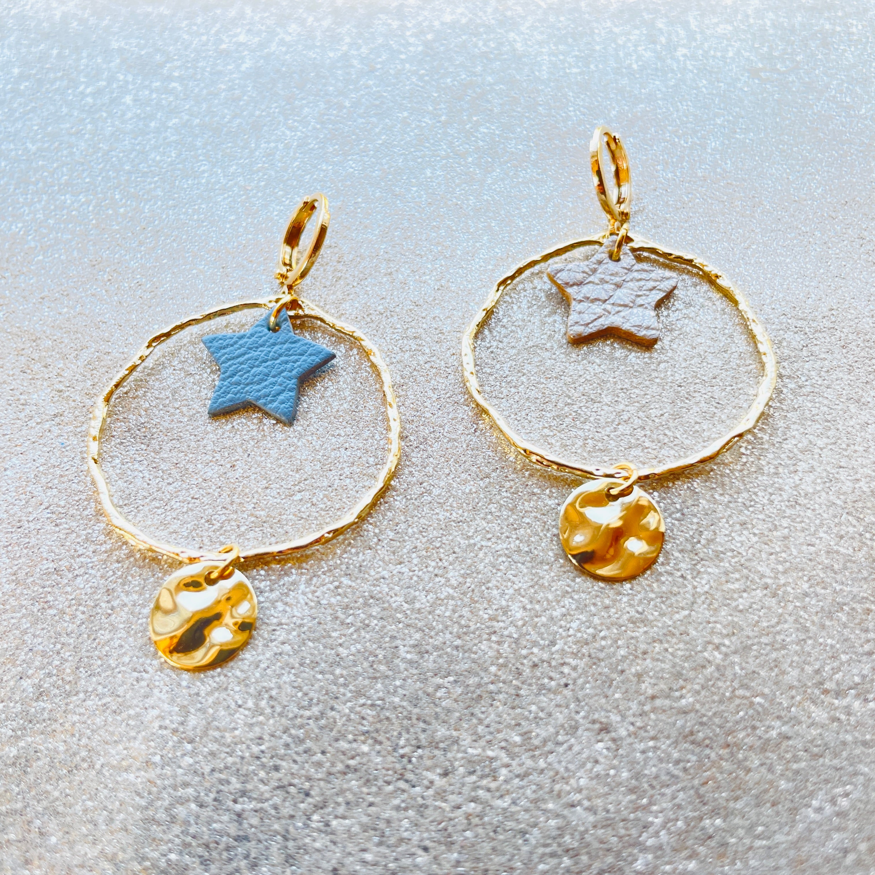 Boucles d'oreilles Nune modèle Naleli dorées à l'or fin de forme créole avec étoile de cuir gris acier et cuir or mat, sur fond cuir beige pailleté