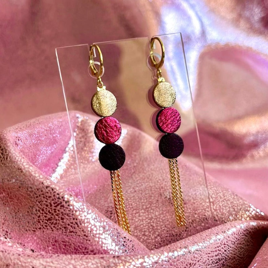 Longues et fines boucles d'oreilles Nune à franges dorées avec 3 petits disques de cuir recyclé cannelle dorée satinée, fuchsia métallisé et violine satinée sur fond cuir rose brillant. Modèle Janis