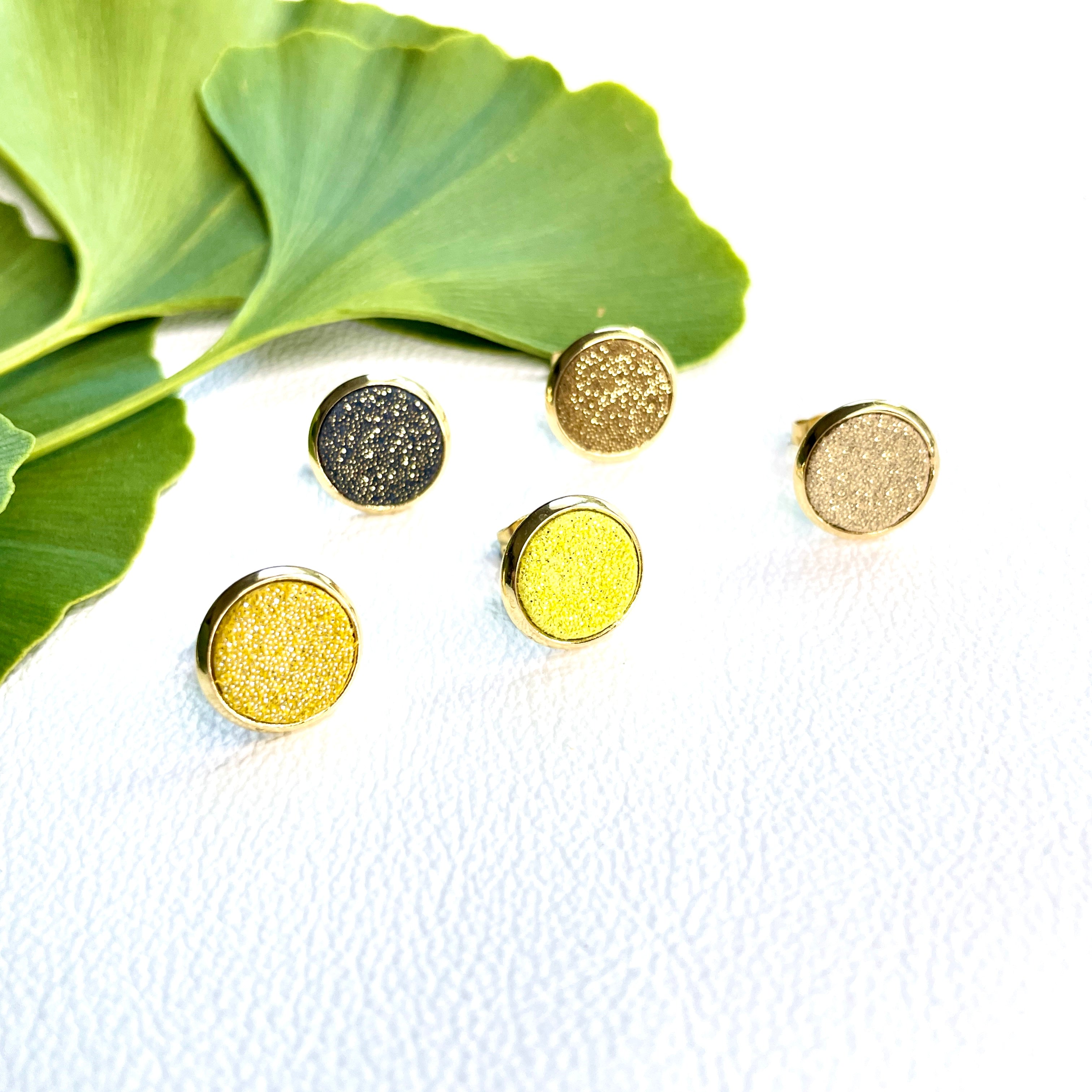 Puces d'oreilles dorées rondes Nune modèle Ilis dans 5 coloris de cuirs pailletés dans les tons jaunes, sur fond blanc avec feuilles de Gingko