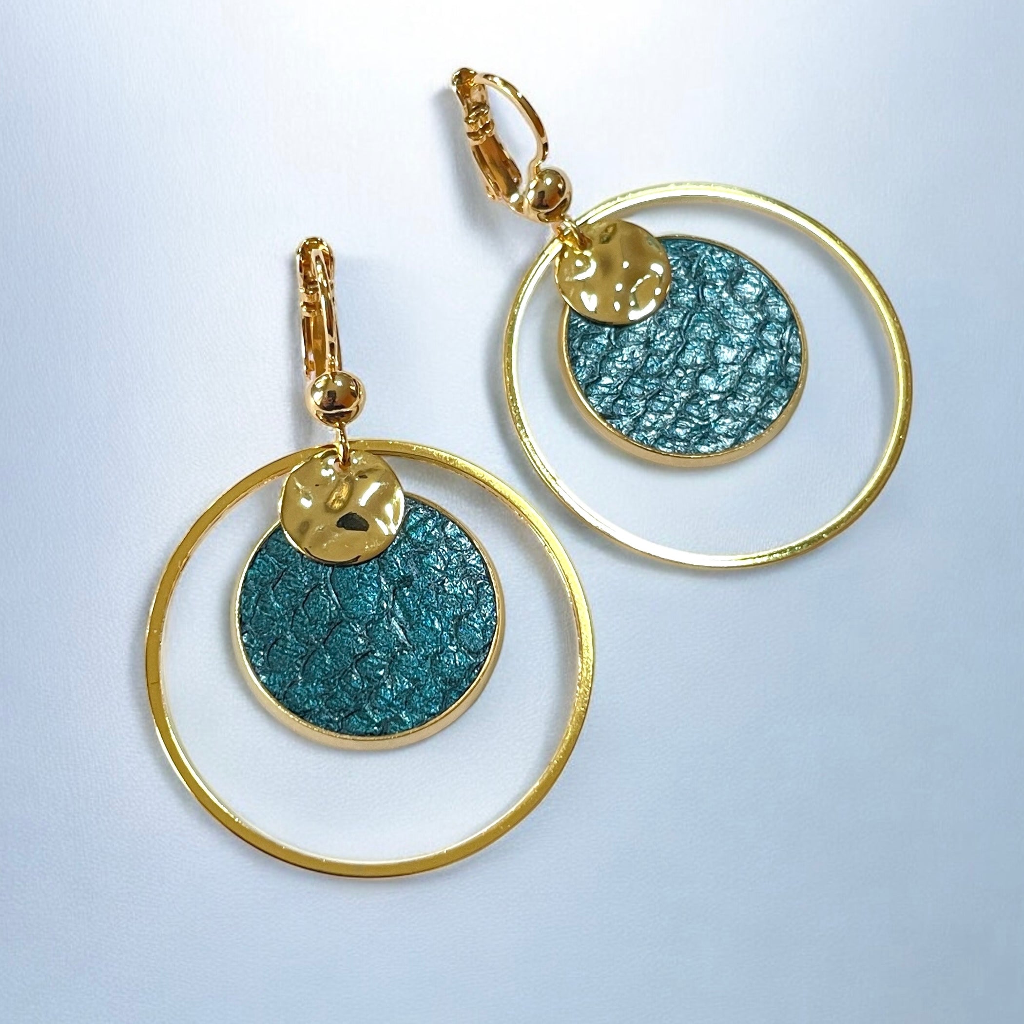 Boucles d'oreilles Arielle de Nune, dorées à l'or fin avec cuir marin bleu paon métallisé, de forme ronde, sur fond cuir bleuté