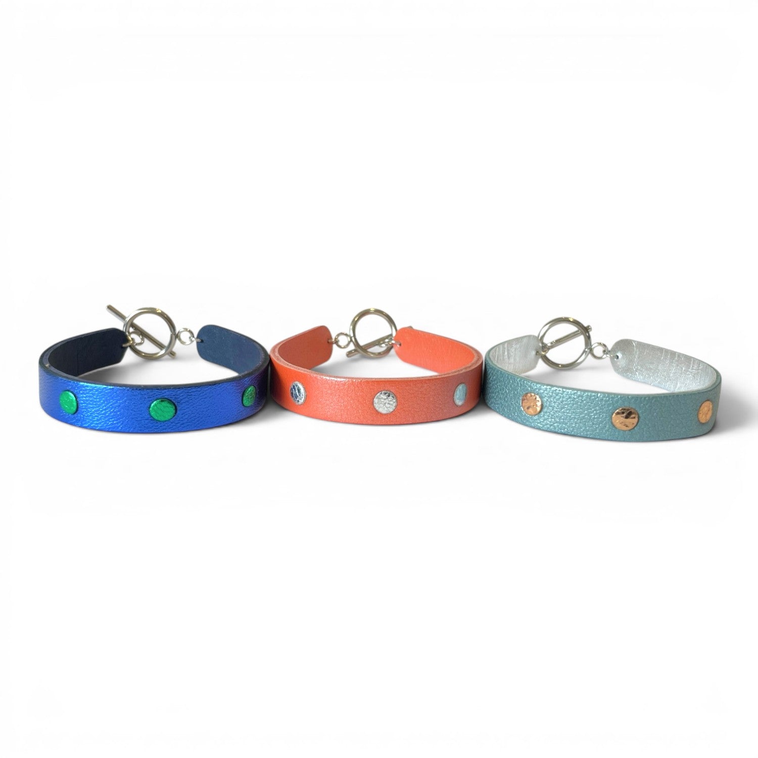 3 Bracelets argentés accolés en cuirs bicolore bleu cobalt métallisé et vert métallisé, corail nacré et argent brillant froissé, et bleu gris nacré et or rosé froissé, empilés les uns sur les autres, sur fond blanc