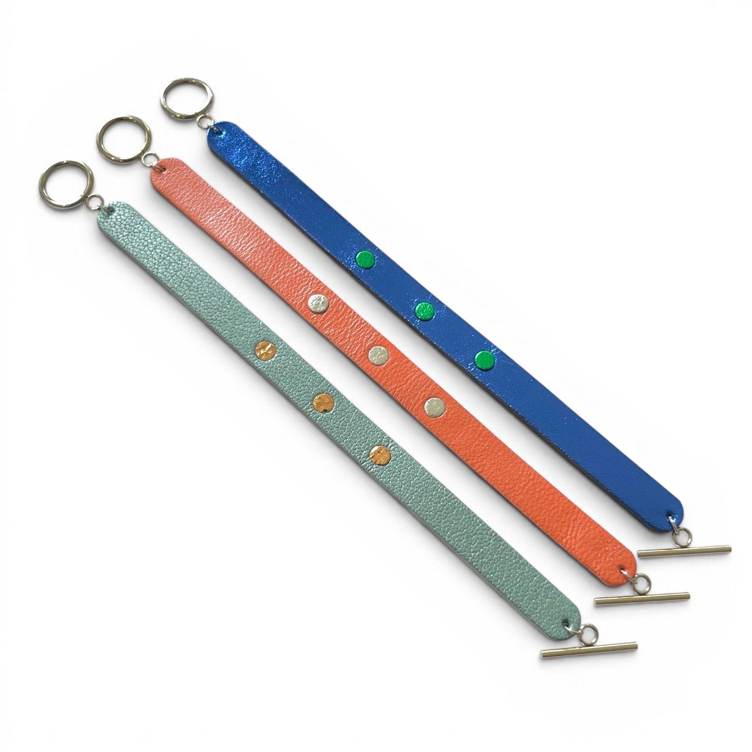 3 Bracelets argentés posés à plat en cuirs bicolore bleu cobalt métallisé et vert métallisé, corail nacré et argent brillant froissé, et bleu gris nacré et or rosé froissé, empilés les uns sur les autres, sur fond blanc