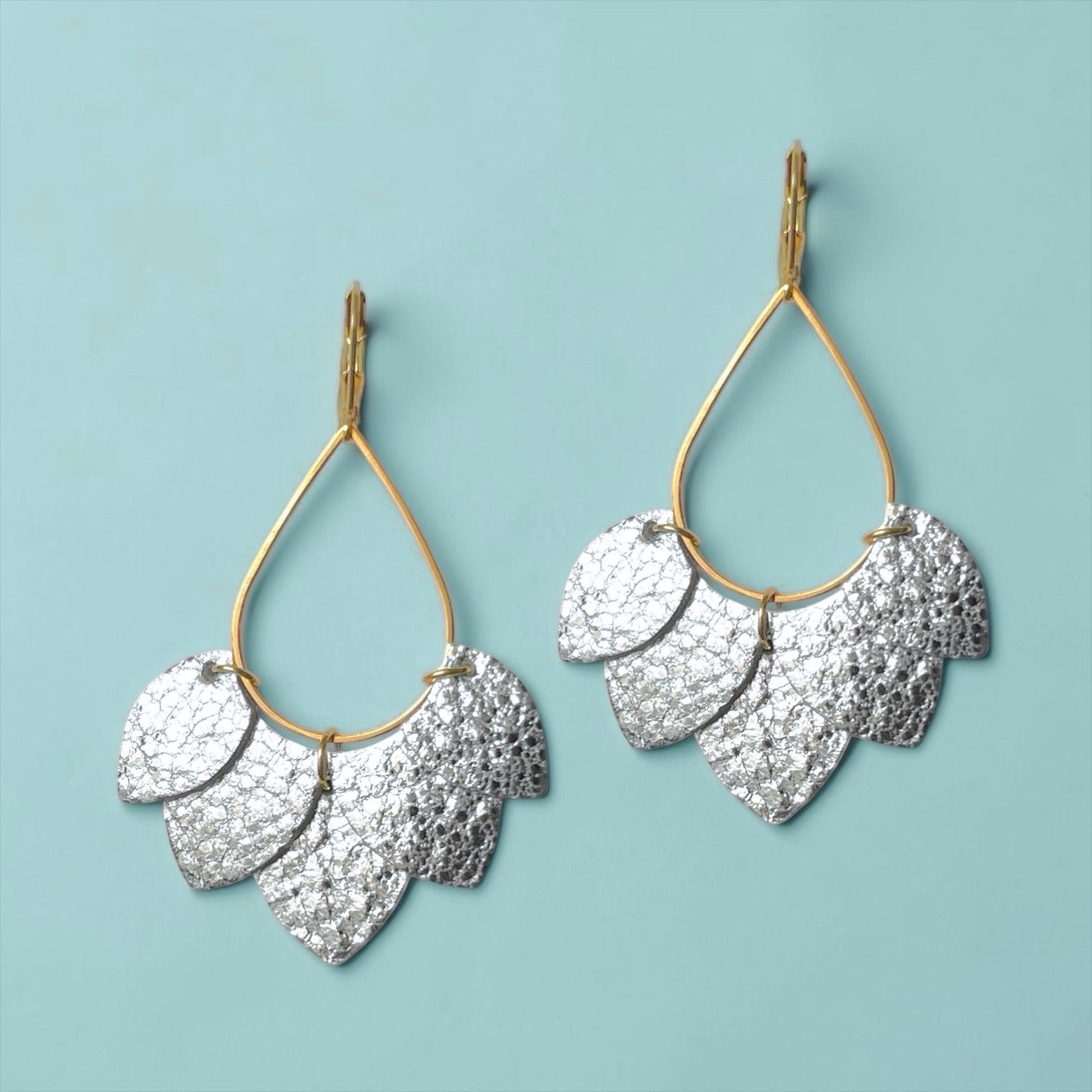 Boucles d'oreilles Nune modèle Aponi dorées à l'or fin avec pétales de cuir argent brillant froissé, sur fond vert