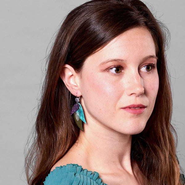 Jeune femme portant les Boucles d'oreilles argentées Nune en forme de plumes, modèle Ara en cuirs violet brillant façon serpent, turquoise métallisé et kaki métallisé sur fond gris