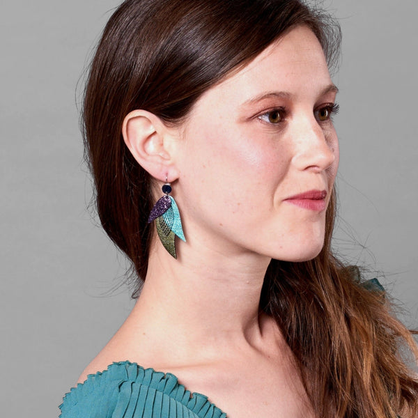 Jeune femme vue de profil portant les Boucles d'oreilles argentées Nune en forme de plumes, modèle Ara en cuirs violet brillant façon serpent, turquoise métallisé et kaki métallisé sur fond gris