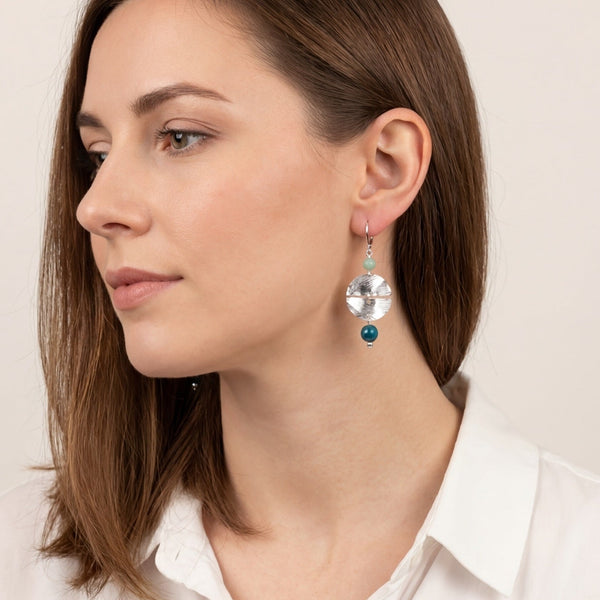 Boucles d'oreilles pendantes de forme ronde avec 2 demi-lunes, en métal martelé argenté et perles d'amazonite et d'apatite bleue, portées à l'oreille par une femme brune