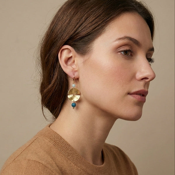 Boucles d'oreilles pendantes de forme ronde avec 2 demi-lunes, en métal martelé doré à l'or fin et perles d'amazonite et d'apatite bleue, portées à l'oreille par une femme brune