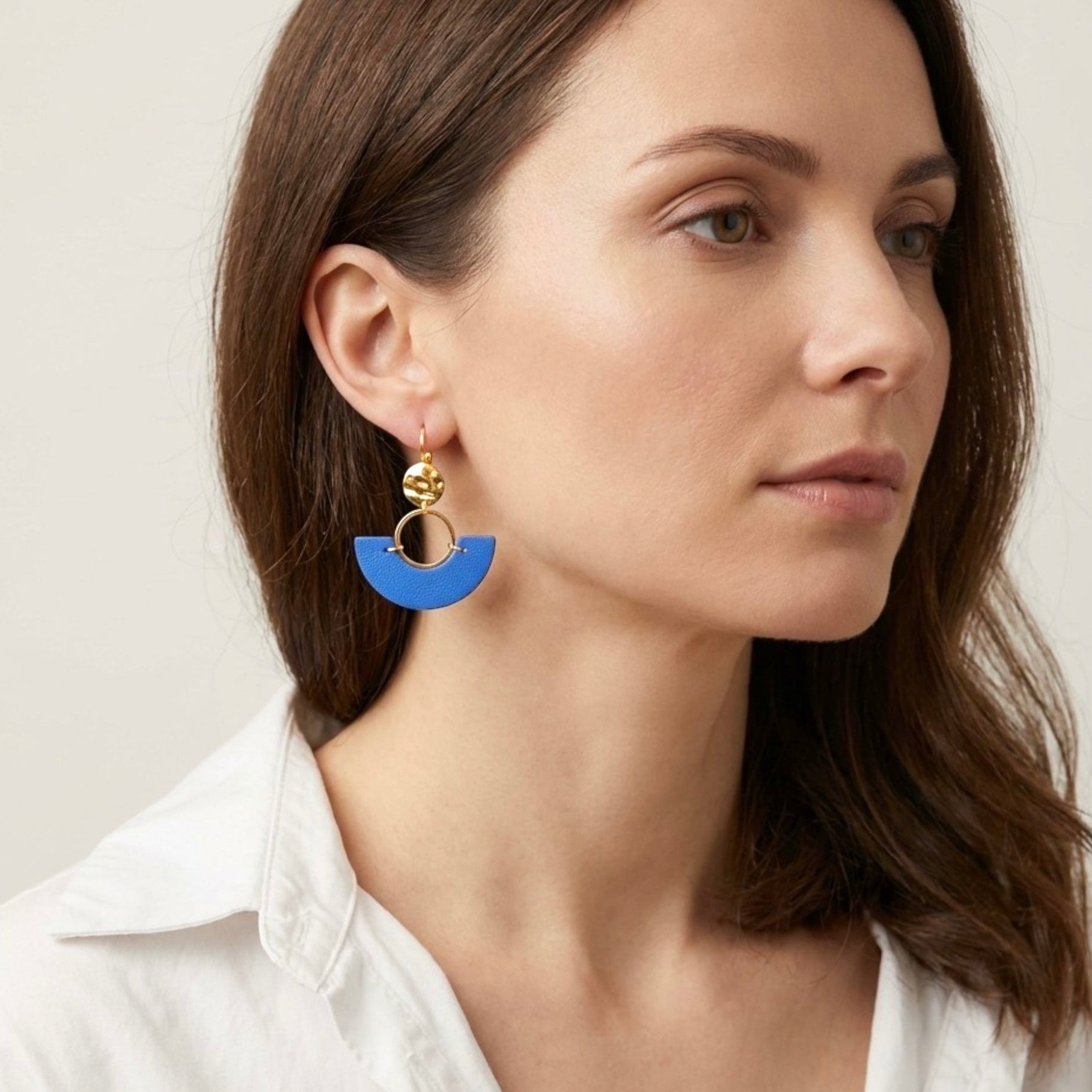 boucles d'oreilles dorées pendantes en forme de demi-lunes en cuir bleu roi, portées à l'oreille par une femme brune