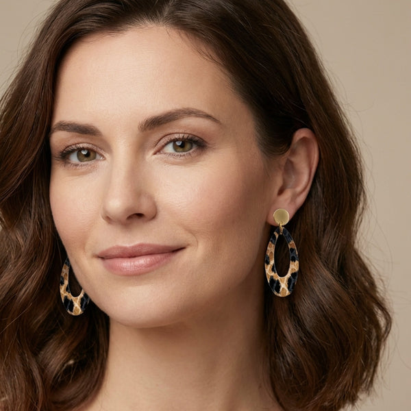 Puces d'oreilles dorées Drys de la marque Nune, avec leurs anneaux ovales en cuir léopard, portées aux oreilles par une femme brune