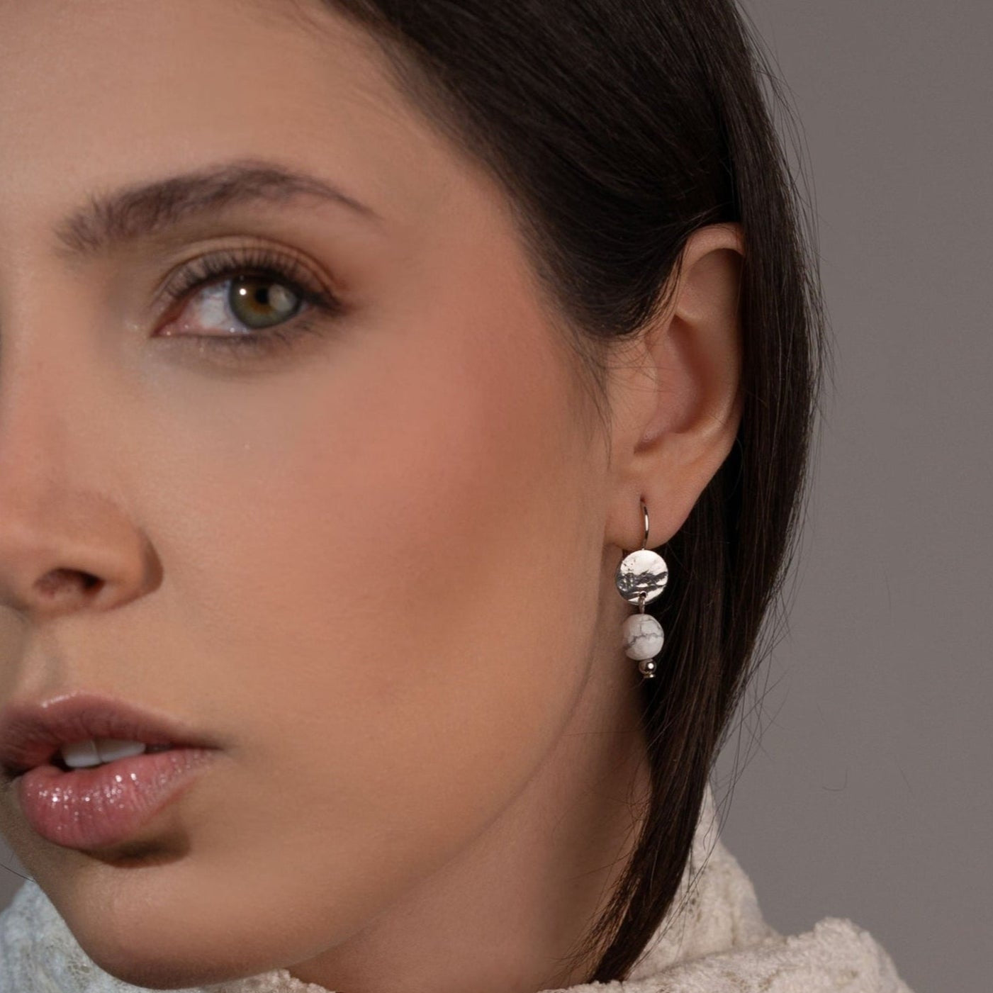 Petites boucles d'oreilles rondes en métal martelé doré à l'or fin avec pendant en perle de howlite blanche, portées à l'oreille par une femme brune