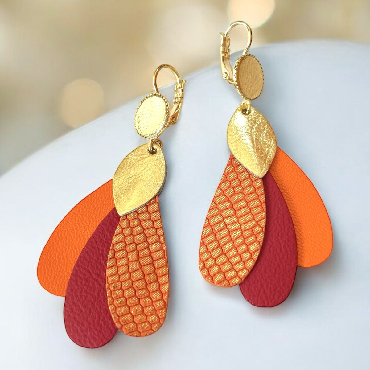 Paire de grandes boucles d'oreilles dorées en forme de plumes de paon, en cuirs recyclés doré, orangé or façon serpent, bordeaux mat et orange, sur fond blanc doré. Modèle Paon de Nune.