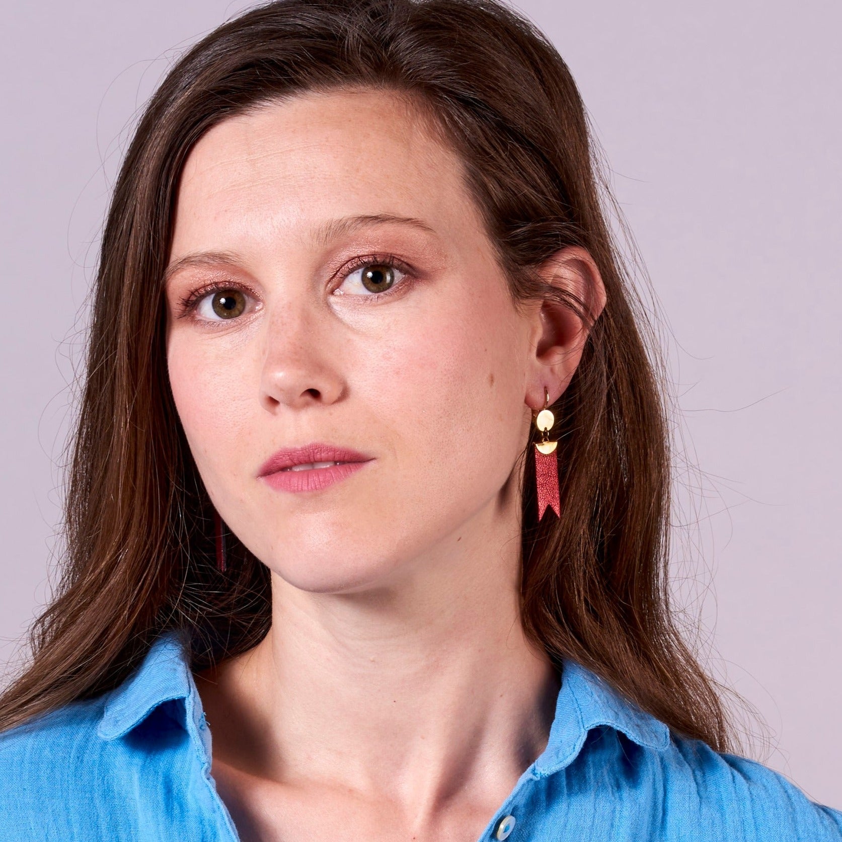 Jeune femme vue de près portant les Boucles d'oreilles dorées Nune modèle Cinta en forme de rubans en cuir rubis métallisé, sur fond gris