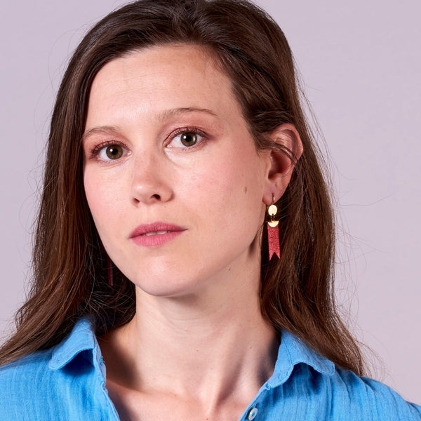 Jeune femme vue de près portant les Boucles d'oreilles dorées Nune modèle Cinta en forme de rubans en cuir rubis métallisé, sur fond gris