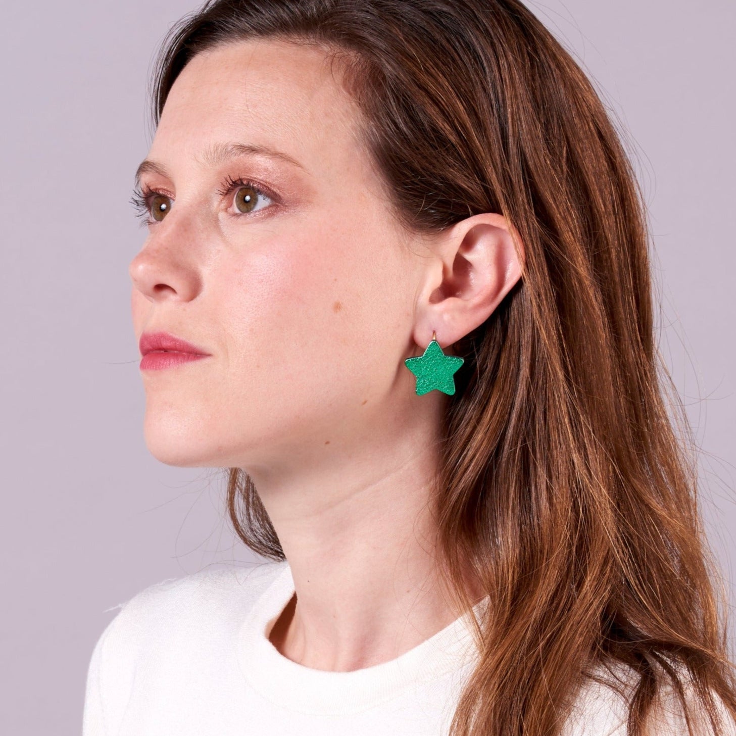 Jeune fille aux longs cheveux bruns avec marinières vue de profil portant les boucles d'oreilles Diva de Nune en forme d'étoile en cuir recyclé vert métallisé, sur mauve