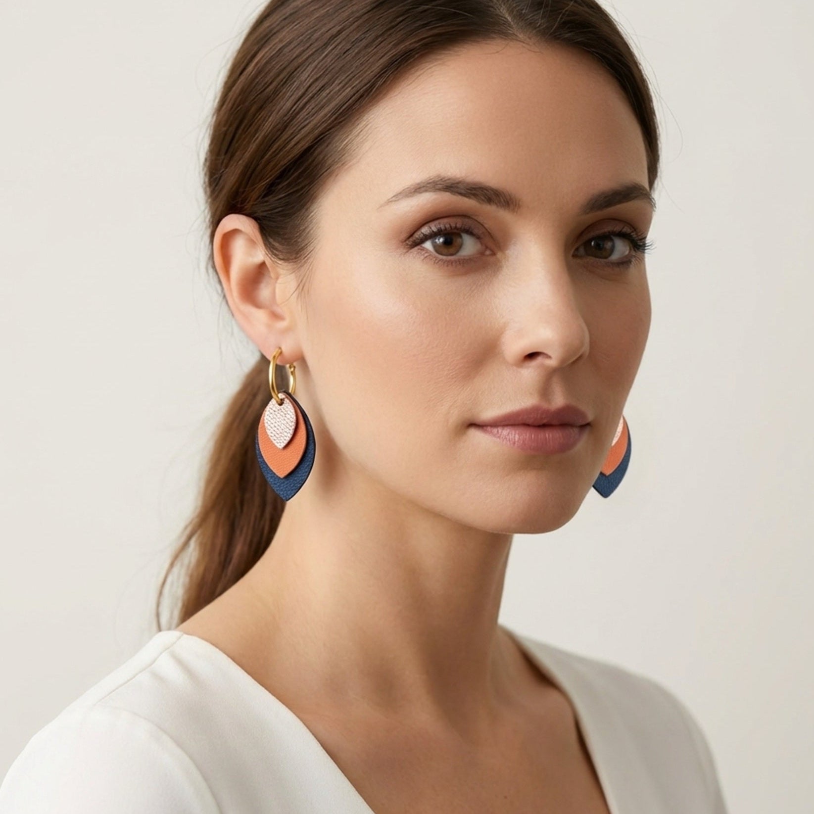 Créoles dorées avec pendentifs modulables en forme de feuilles en cuirs rose pâle métallisé, saumon clair et bleu marine, portées à l'oreille par une femme brune