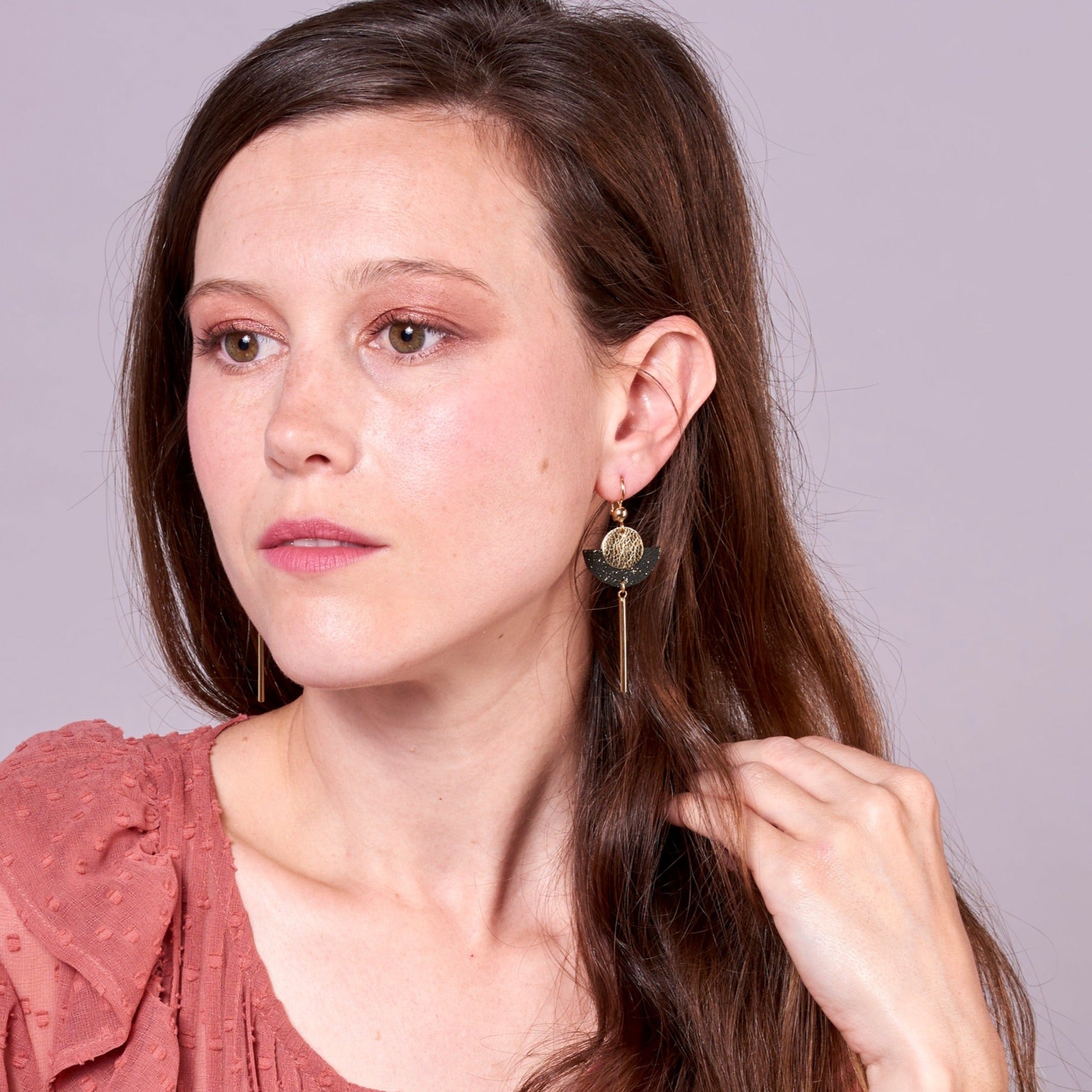 Jeune femme vue de face portant les Longues boucles d'oreilles dorées en forme de demi-lune en cuirs doré et noir pailleté or recyclé, sur fond mauve