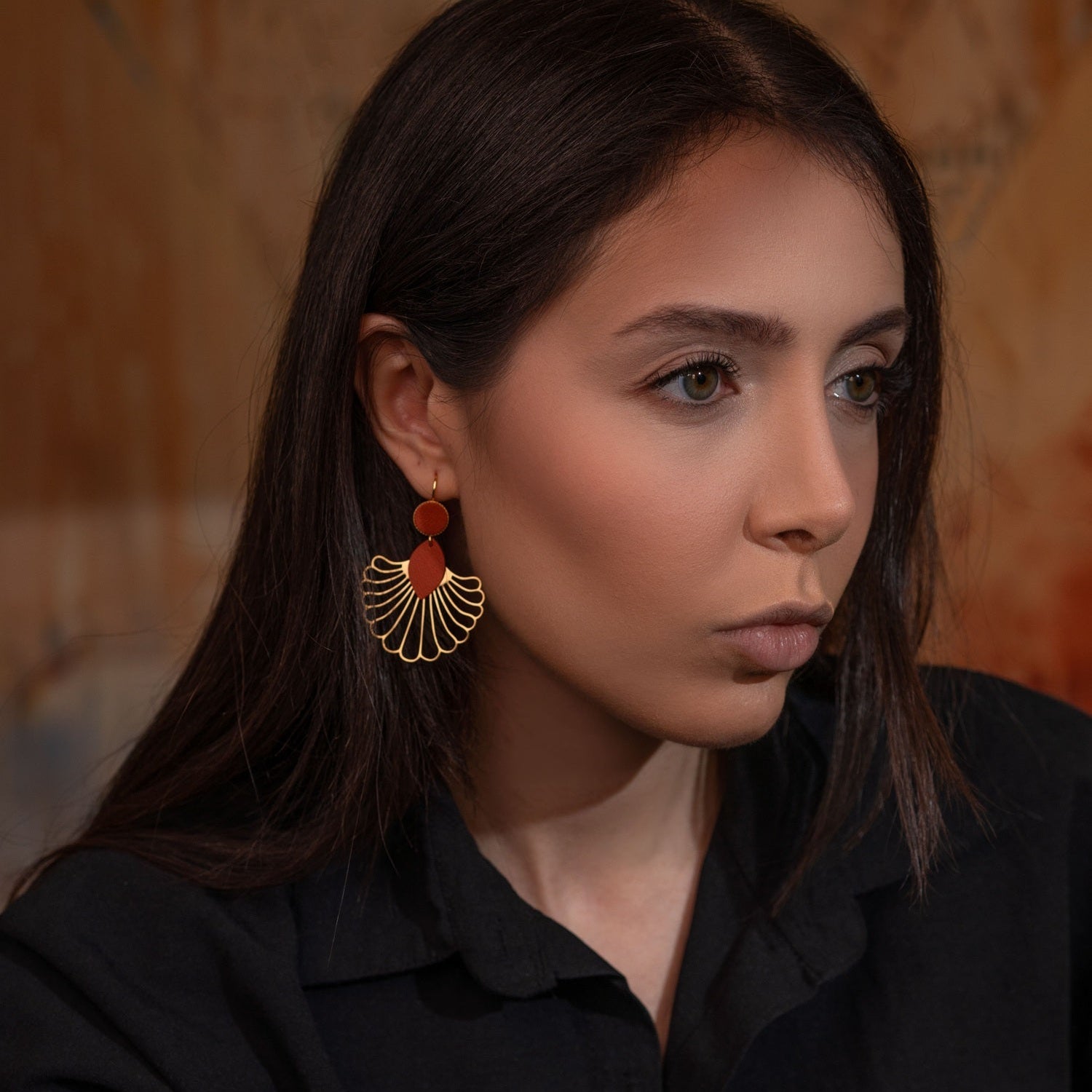 Grandes boucles d'oreilles dorées en forme d'éventail style art déco, avec empiècements de cuir terracotta, portées à l'oreille par une femme brune