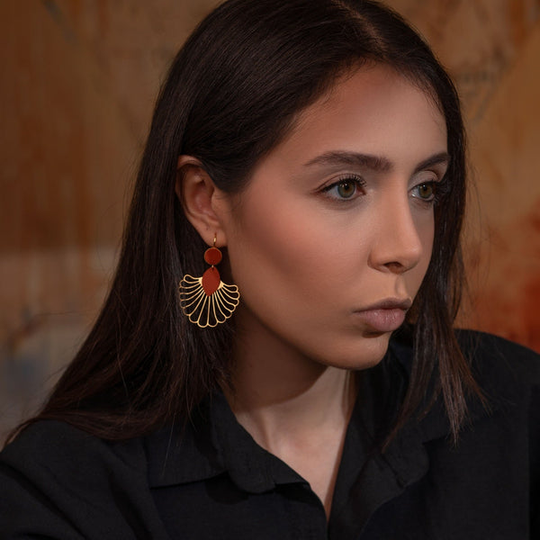 Grandes boucles d'oreilles dorées en forme d'éventail style art déco, avec empiècements de cuir terracotta, portées à l'oreille par une femme brune