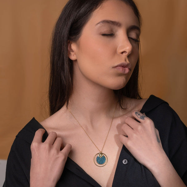 Fin collier doré avec pendentif rond en anneau avec une pièce de cuir marin bleu paon métallisé,  porté par une femme brune