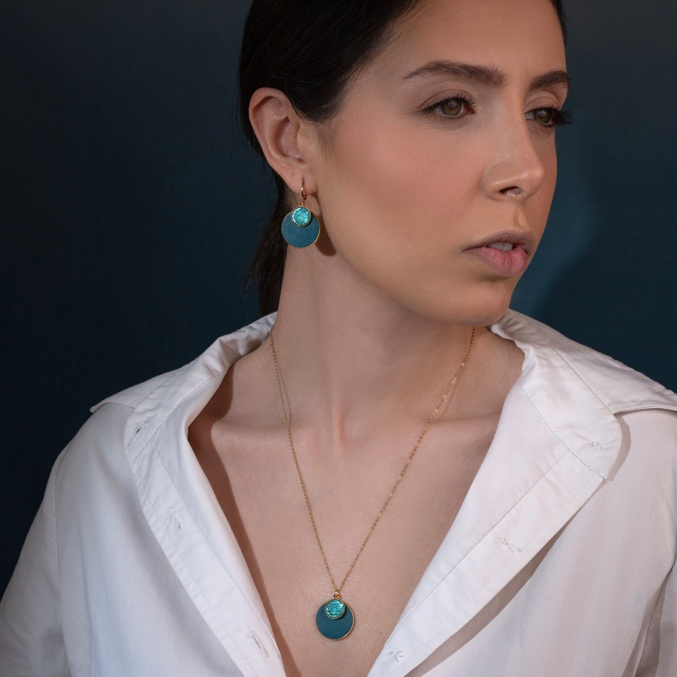 Collier Bulles Nune, court doré avec pendentif rond composé de doubles sertis de cuirs vert canard et vert émeraude brillant intense, et boucles d'oreilles assorties, portées par une femme brune