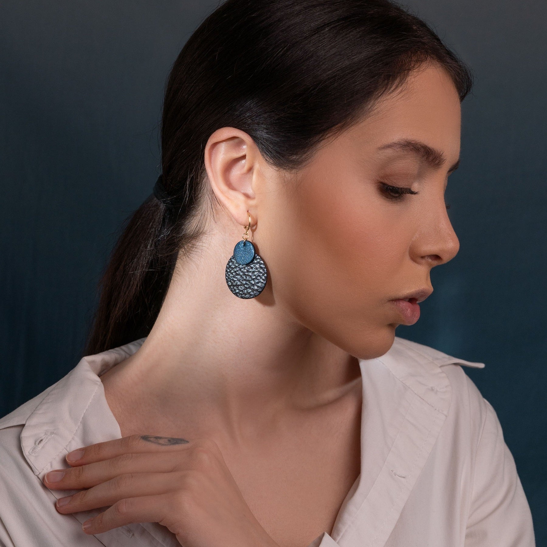 boucles d'oreilles Chica pendantes en duo de disques bicolores bleu nuit métallisé et bleu nuit grainé métallisé, portées à l'oreille par une femme brune