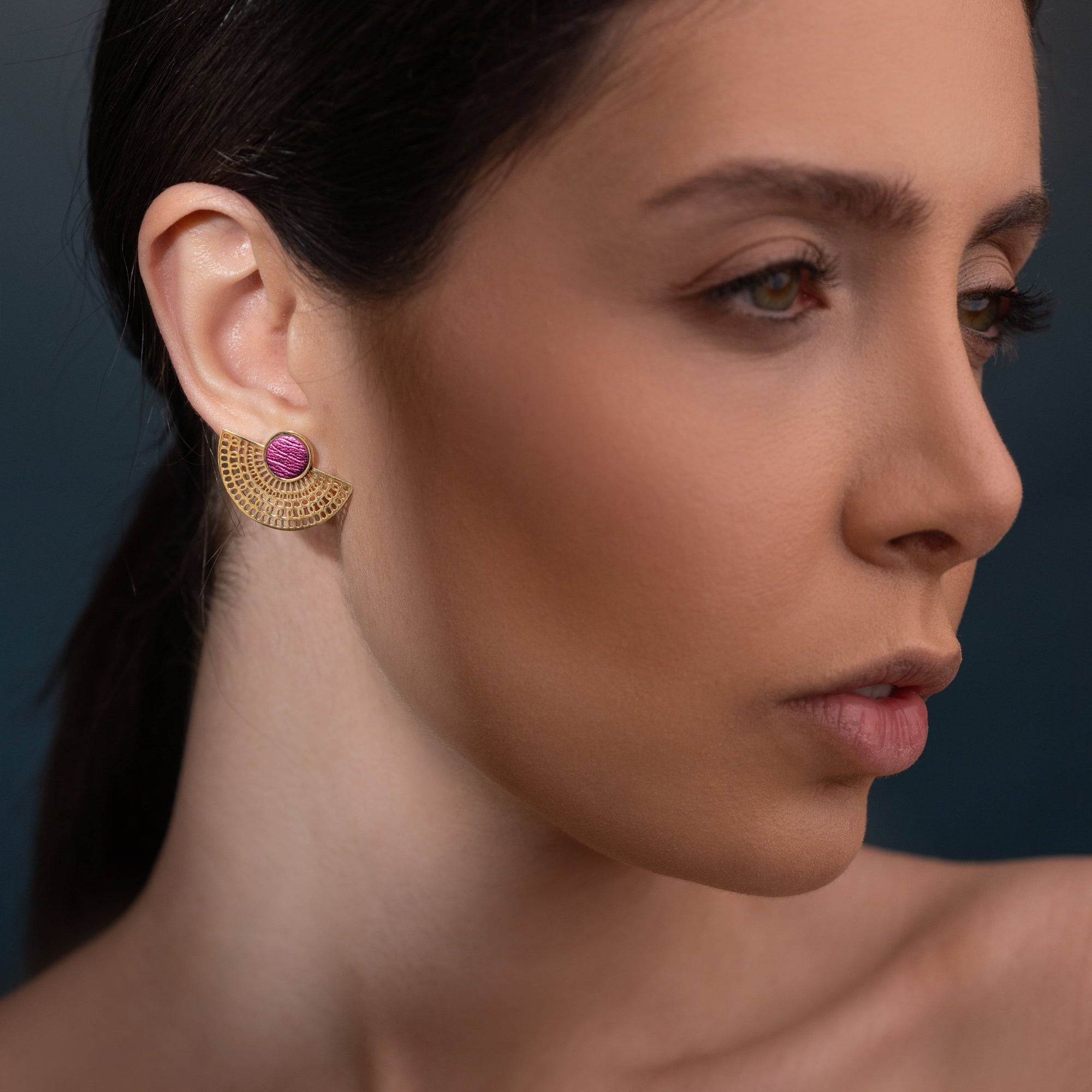 boucles d'oreilles puces dorées Nune modèle Amphea en cuir rose fushia métallisé, portées à l'oreille par une femme brune vue de près