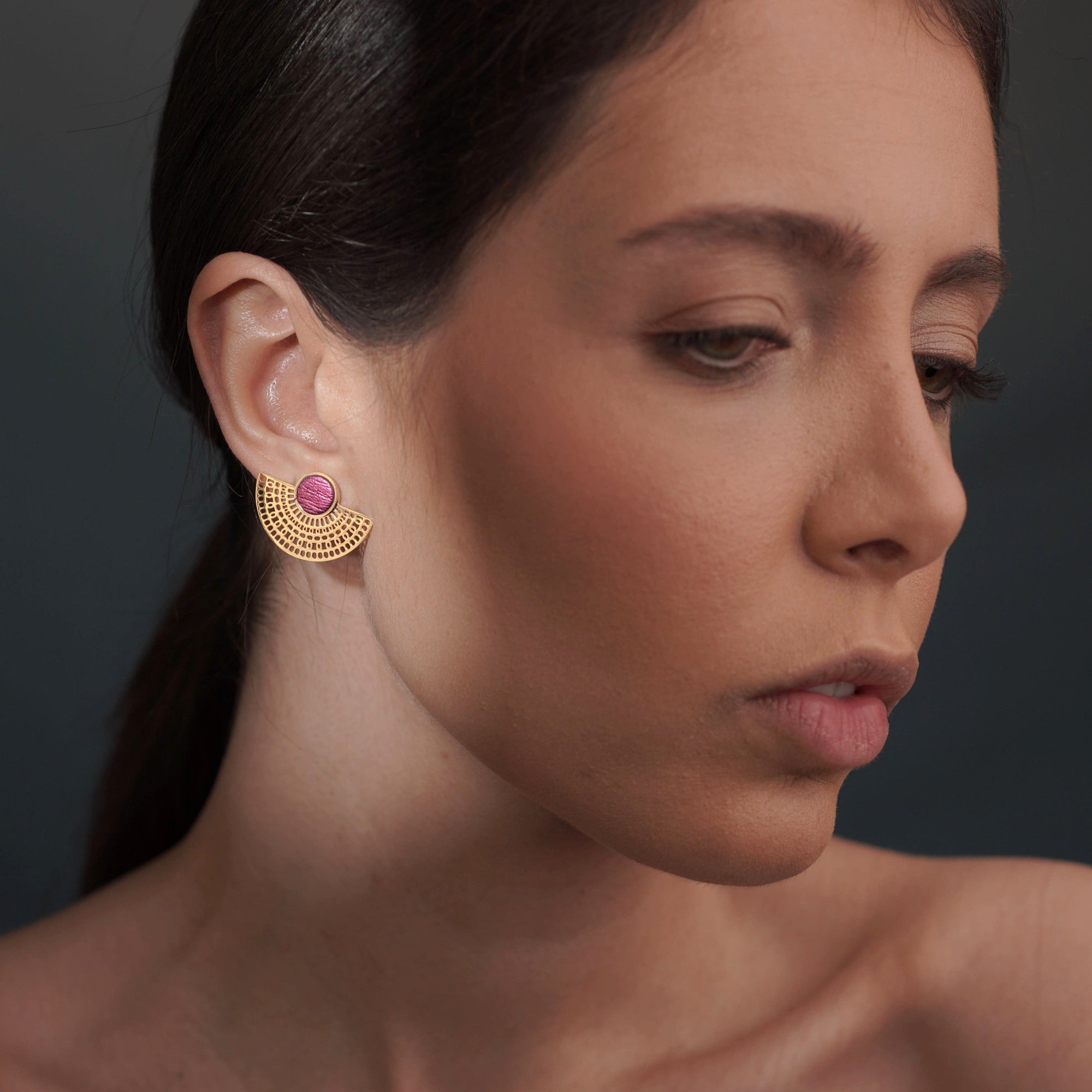 boucles d'oreilles puces dorées Nune modèle Amphea en cuir rose fushia métallisé, portées à l'oreille par une femme brune