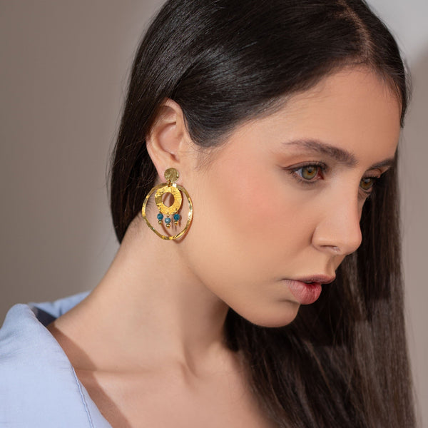 Grandes boucles d'oreilles puces créoles dorées pendantes longues rondes avec double anneau et petites perles en apatite bleue, portées à l'oreille par une femme brune