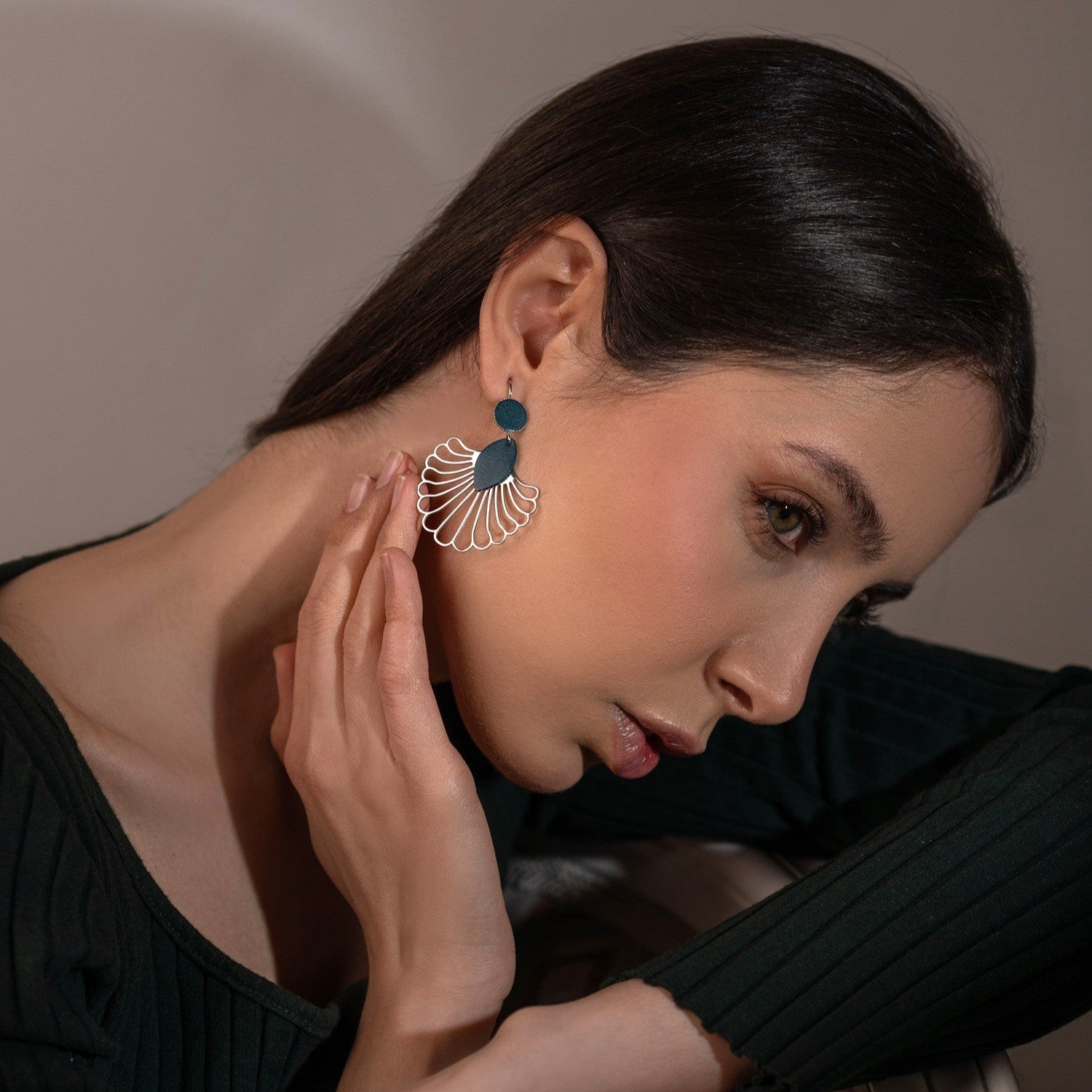 Grandes boucles d'oreilles argentées en forme d'éventail style art déco, avec empiècements de cuir vert canard, portées à l'oreille par une femme brune