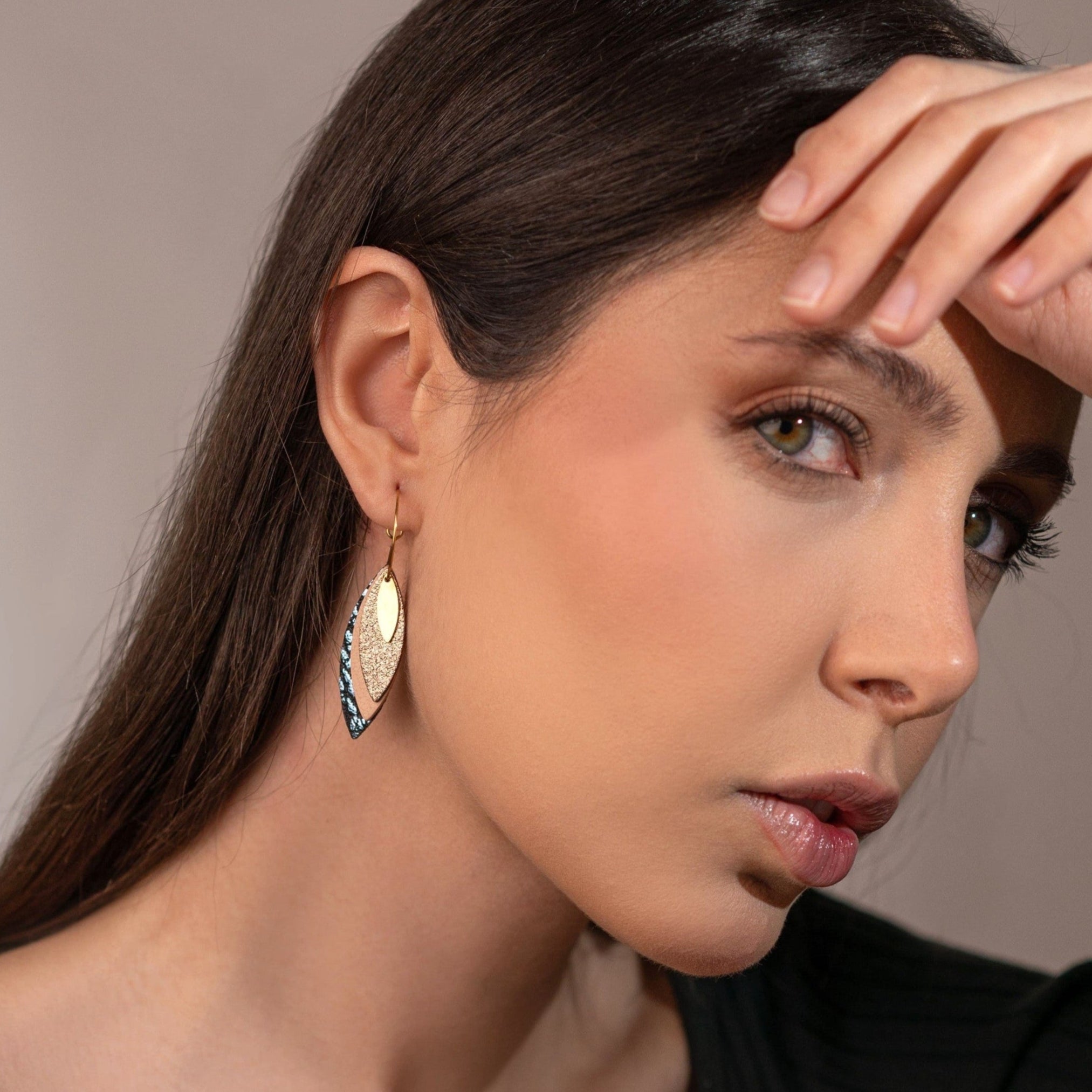 Boucles d'oreilles créoles dorées Nune modèle Piume en cuirs cannelle dorée satinée, rose pâle et bleu glacier métallisé grainé, portées à l'oreille par une femme brune