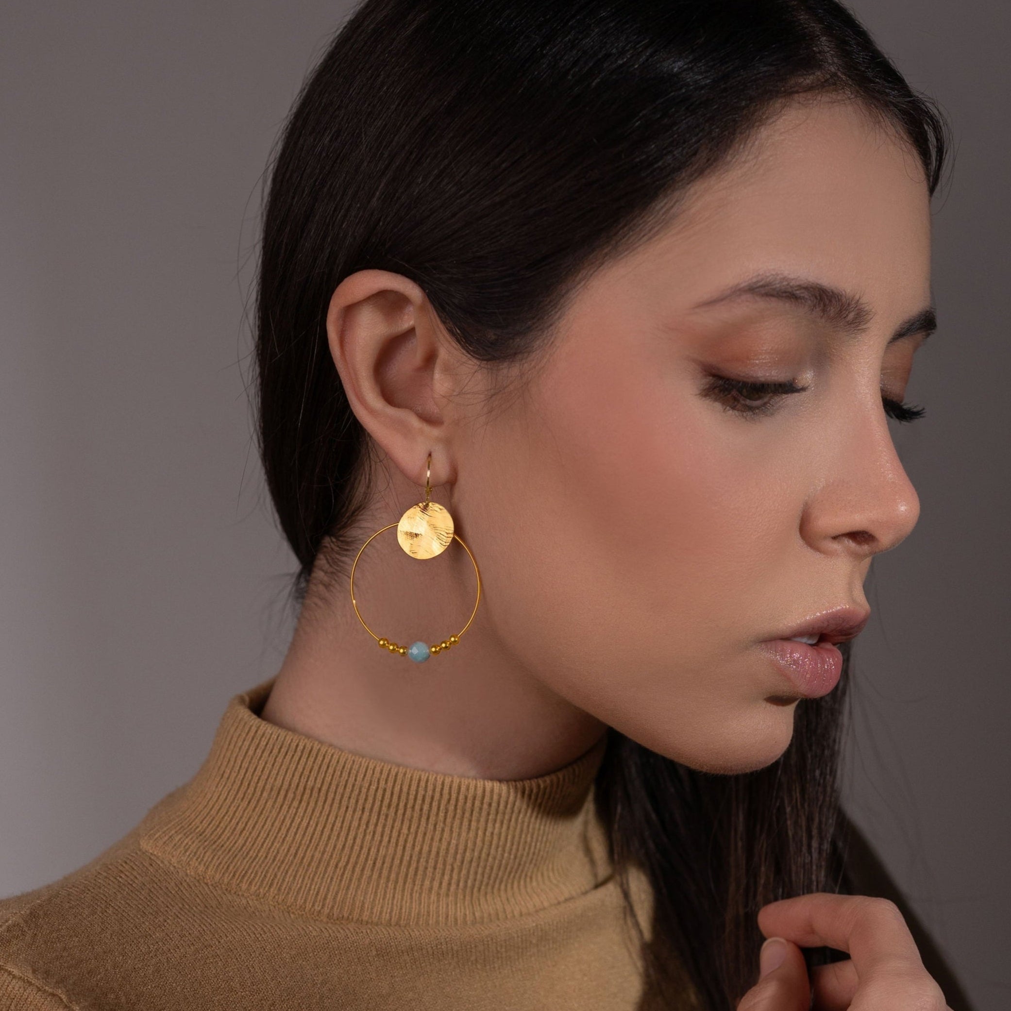 Boucles d'oreilles créoles dorées pendantes longues, style créoles, avec sequin et perle en amazonite verte, portées à l'oreille par une femme brune