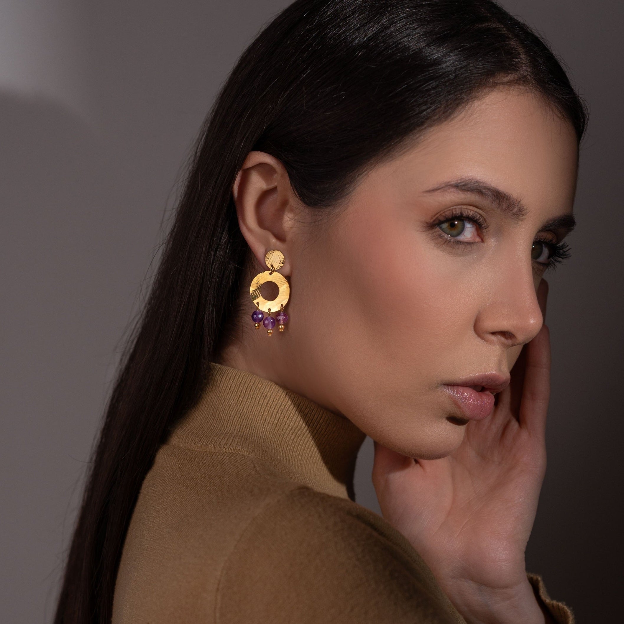 Petites boucles d'oreilles créoles à puces de forme ronde en métal martelé doré à l'or fin et pierres naturelles améthystes, portées à l'oreille par une femme brune