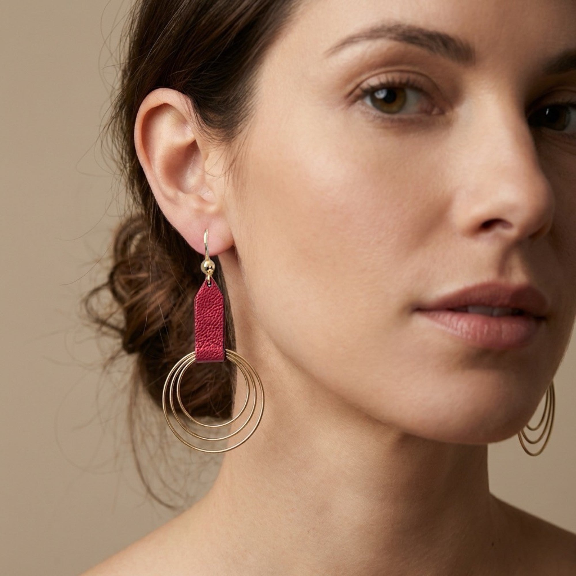Grandes boucles d'oreilles dorées avec pendentif en anneaux concentriques attaché par un ruban de cuir recyclé rubis métallisé, portée aux oreilles. 