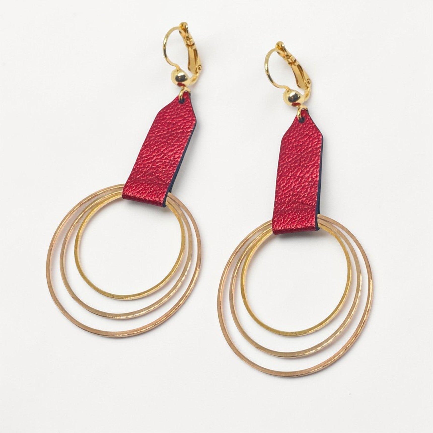 Grandes boucles d'oreilles dorées avec pendentifs en anneaux concentriques attachés par des rubans de cuir recyclé rubis métallisé, sur fond blanc. Modèle Dizzo