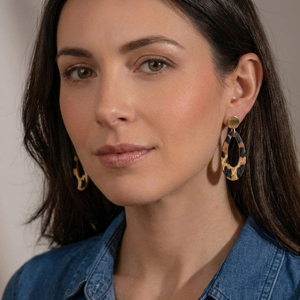 Puces d'oreilles dorées Drys de la marque Nune, avec leurs anneaux ovales en cuir léopard, portées aux oreilles par une femme brune