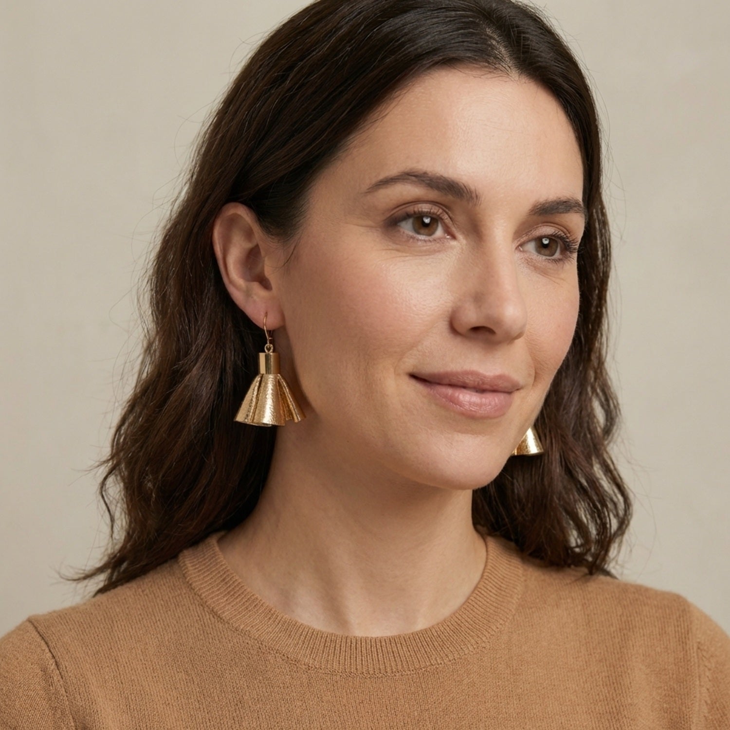 Boucles d'oreilles dorées en pompons frou frou de cuir doré, portées par une femme brune avec pull camel