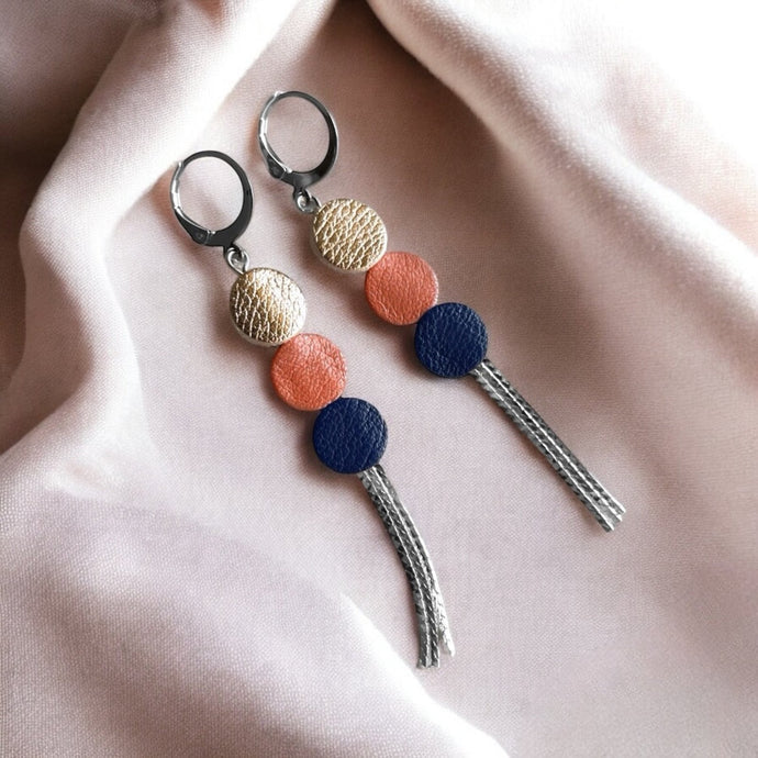 Longues et fines boucles d'oreilles à franges argentées avec 3 petits disques de cuir recyclé rose pâle métallisé, corail nacré et bleu marine satiné sur un tissu beige