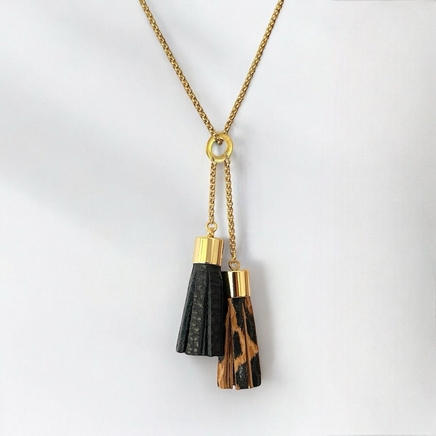 Fin sautoir doré avec pendentif en duo de pompons en cuirs noir grainé et léopard, sur fond blanc