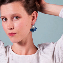 Charger l&#39;image dans la galerie, Boucles d&#39;oreilles argentées Nune modèle Sakura en pétales de cuir bleu nuit métallisé, portées par une jeune femme sur fond bleu gris
