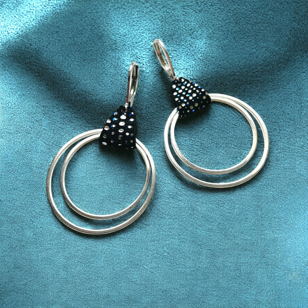 Créoles argentées du modèle Vega de la marque Nune, avec une touche de cuir noir à pois argentés et bleus, sur fond bleu