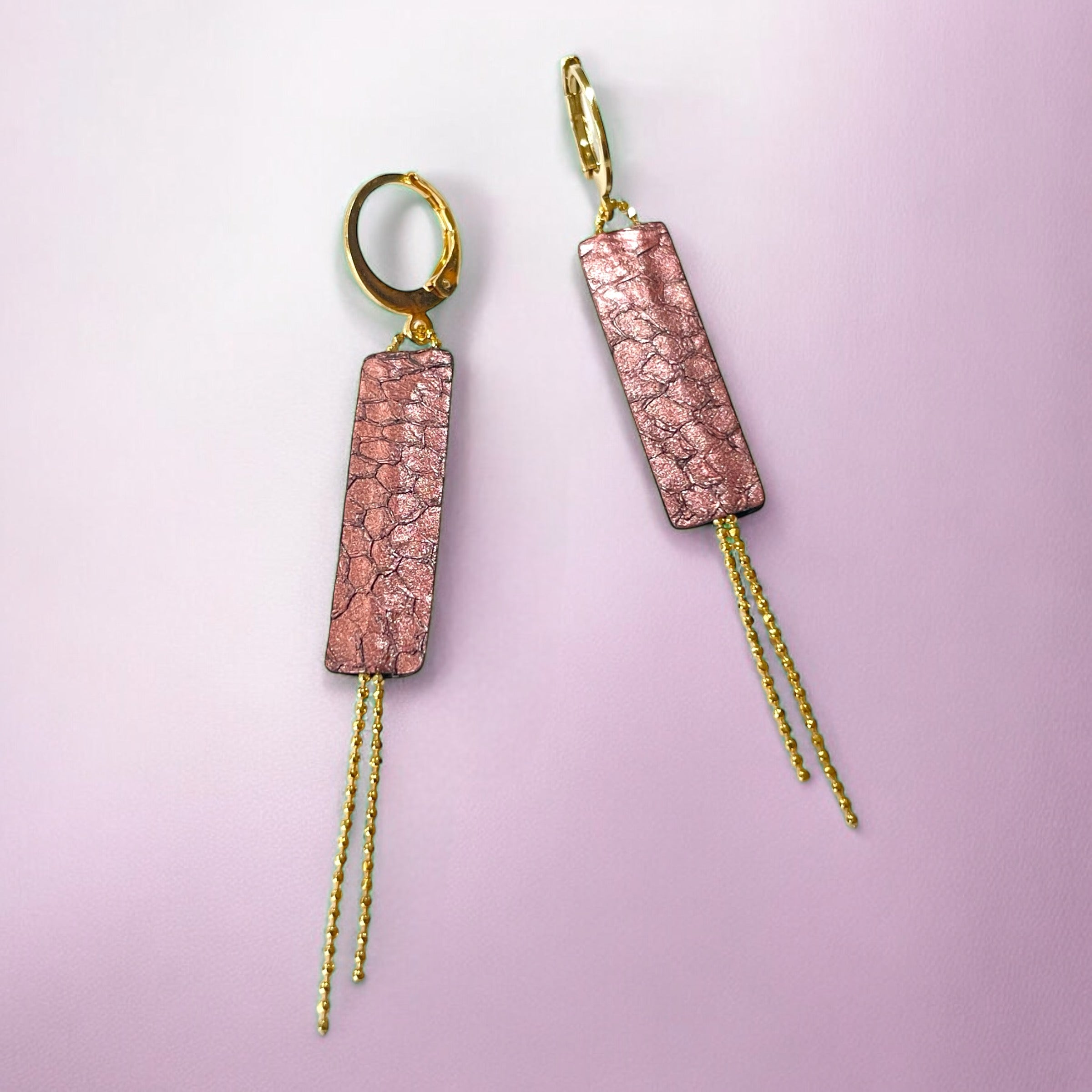 Boucles d'oreilles Effinis de Nune, dorées à l'or fin avec cuir marin, cuir de poisson saumon veux rose métallisé, de forme effilée avec chaînettes dorées pendantes, sur fond rose